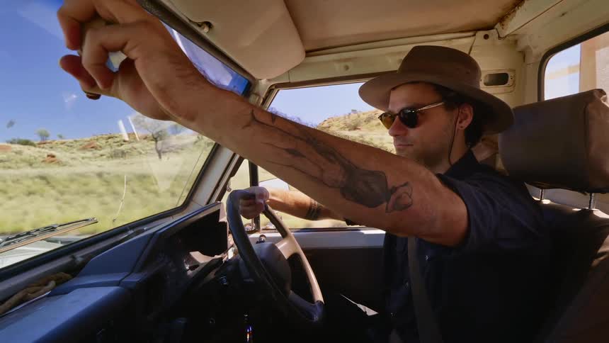 A man wearing sunglasses and a sun hat drives a vehicle through the wilderness