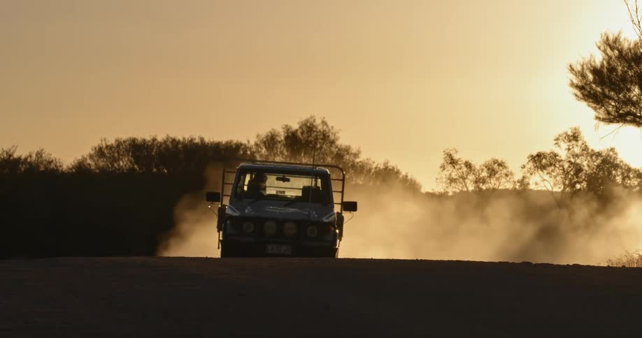 A truck speeds and kicks up dust on a rural dirt road at sunset