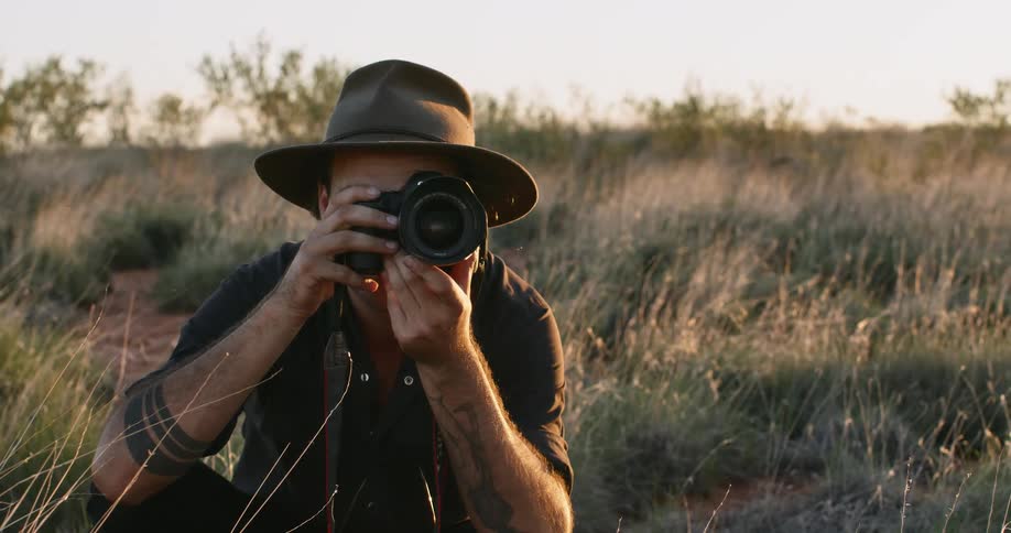 A photographer kneels in the grass with his camera raised to his face to take a photograph