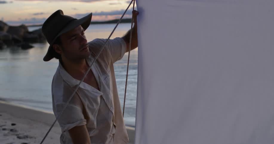 Man wearing a hat sets up a tent on the sand in the beach at sunset