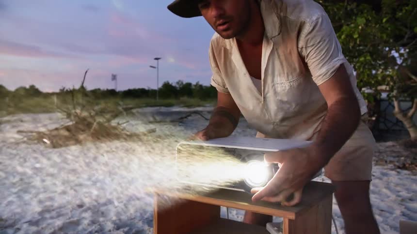 A man sets up a projector on a table on a sandy beach wearing dirty clothing