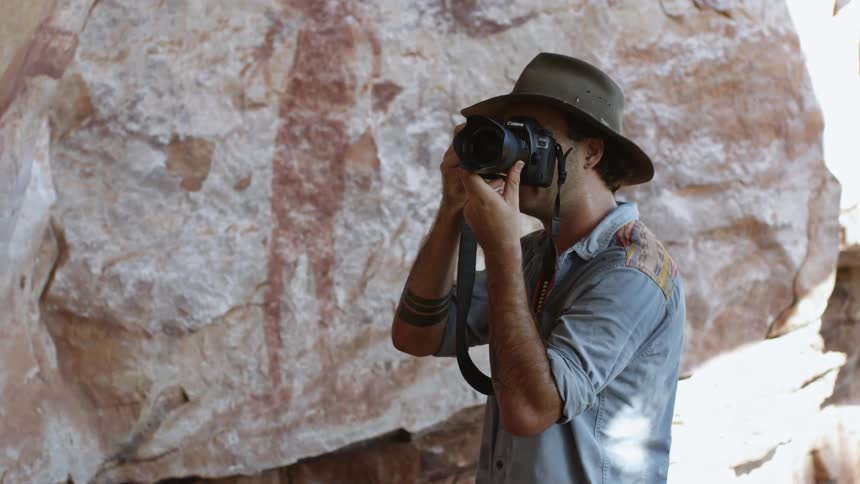 Man wearing a cowboy hat stands near a large rock as he takes a photo with a camera