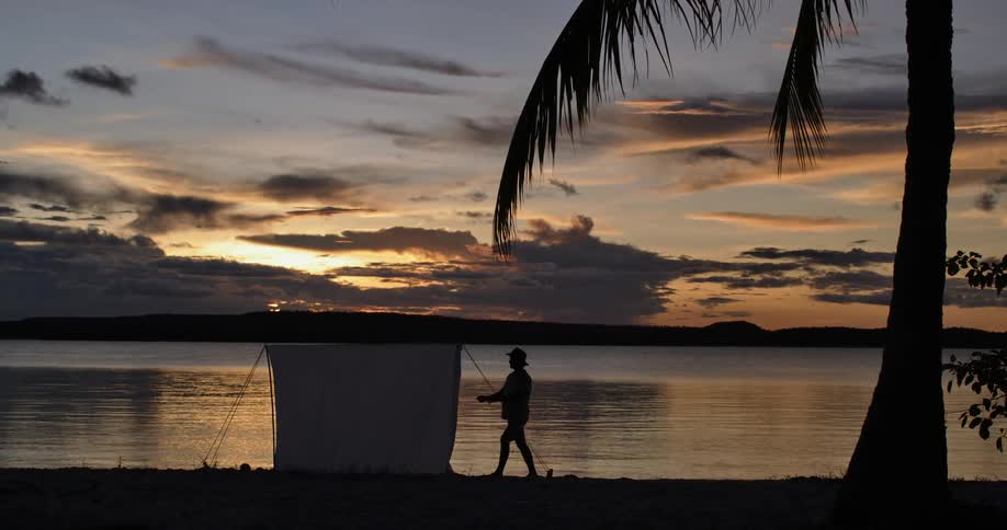 The silhouette of a person can be seen setting up a tent on the beach as the sun is setting