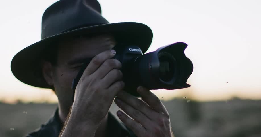 Man wearing a hat takes pictures with a camera surrounded by flying insects at sunset