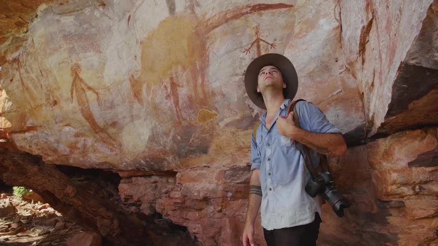 Man wearing a hat and with a camera hanging at his side looks around at the large boulders