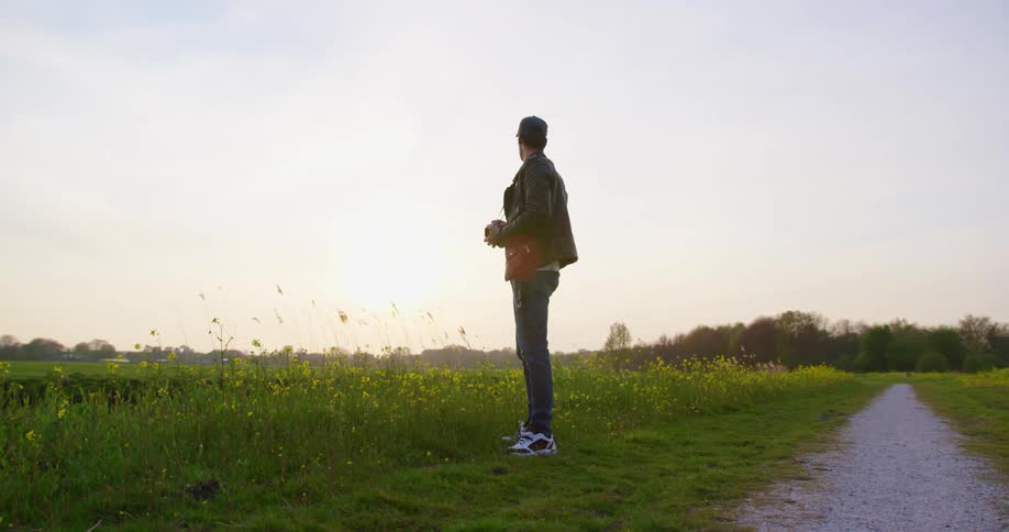 A man with a hat jacket and camera standing on grass next to a pathway