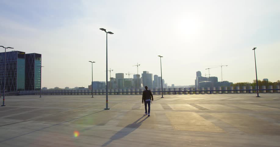A man in a leather jacket walks along a boardwalk in a city on a sunny day