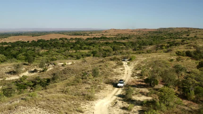 Vehicles drive along dirt road through fields and meadows near hills