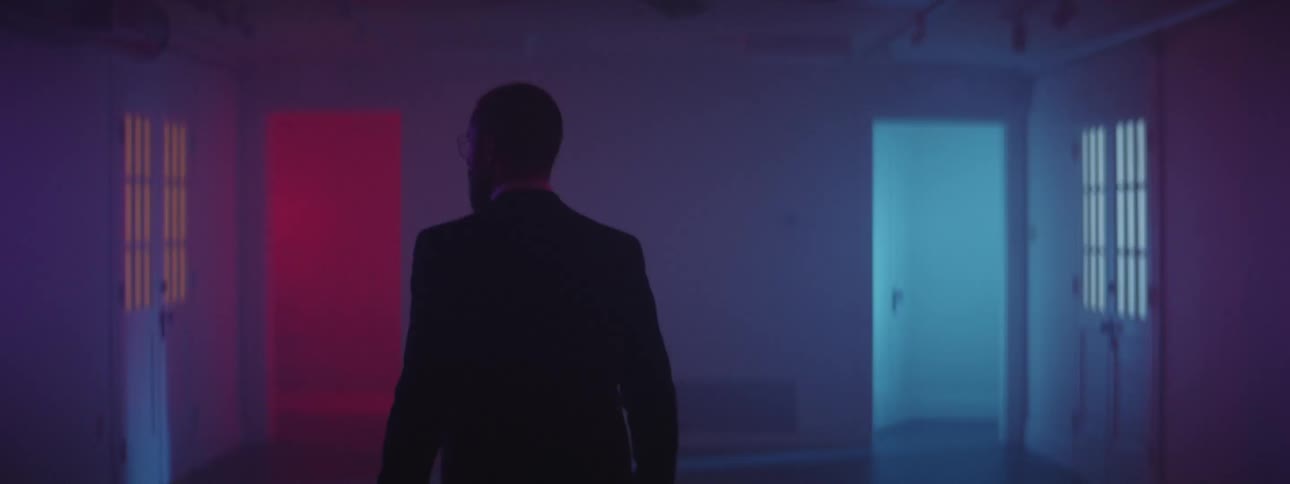 A man in a suit walks through an empty room illuminated by purple light towards a wall with a doorway illuminated by red light and a door illuminated by blue light
