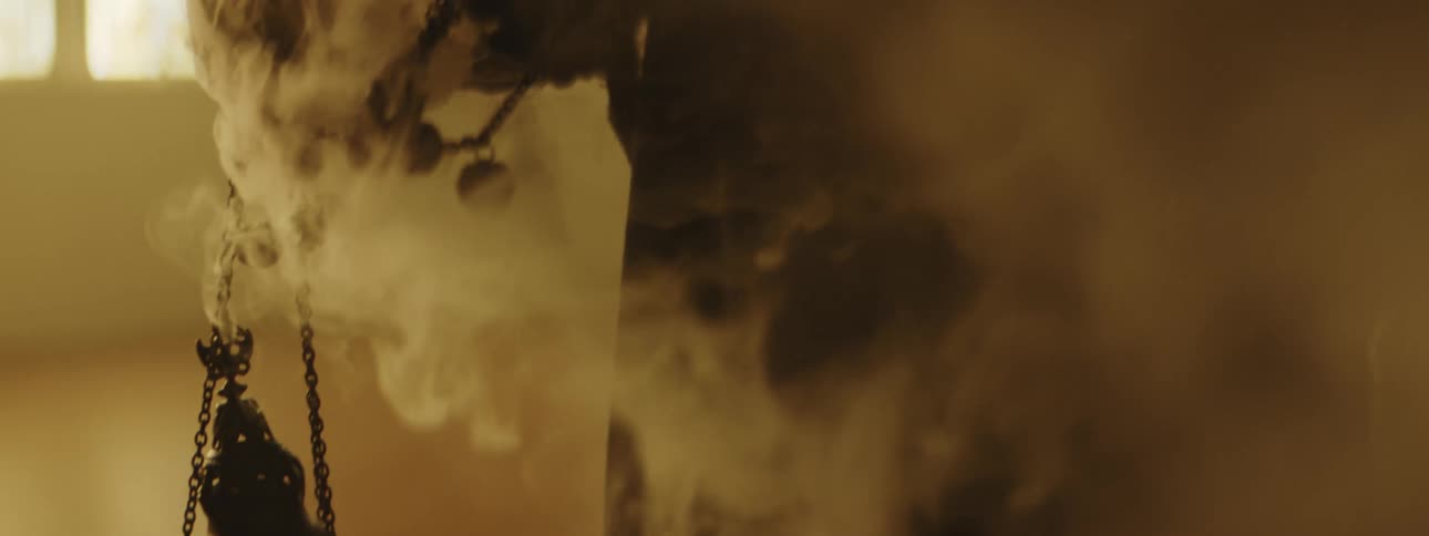 Smoke swirls around a hooded figure indoors