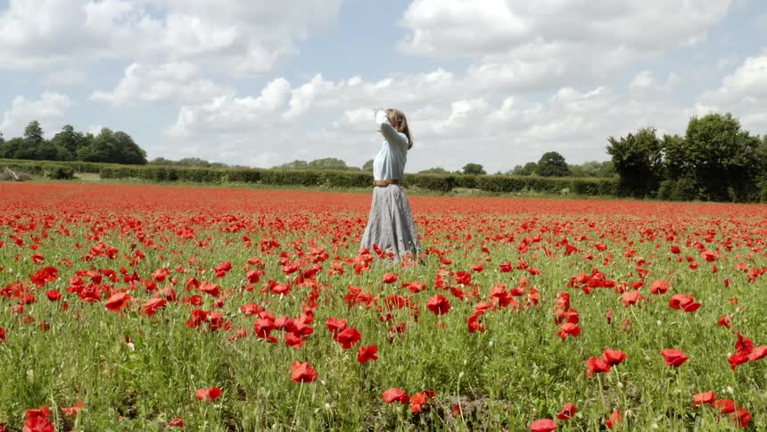 Woman stands in a field of flowers and shields her eyes from the sun as a car goes by in the distance