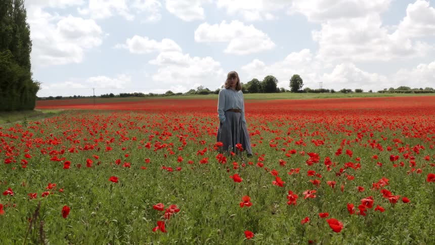 A woman in old fashioned clothing looks into the camera while standing in a field of red flowers