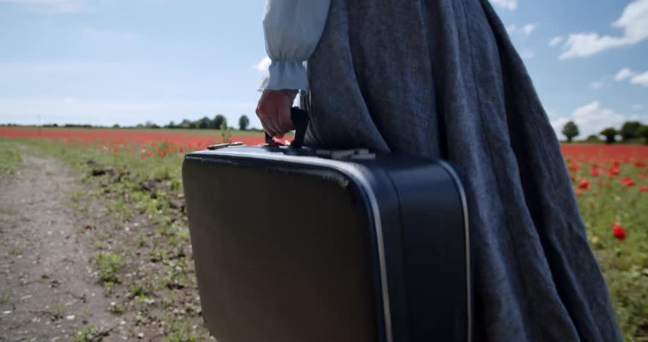 A caucasian female walking along a road carrying a suitcase
