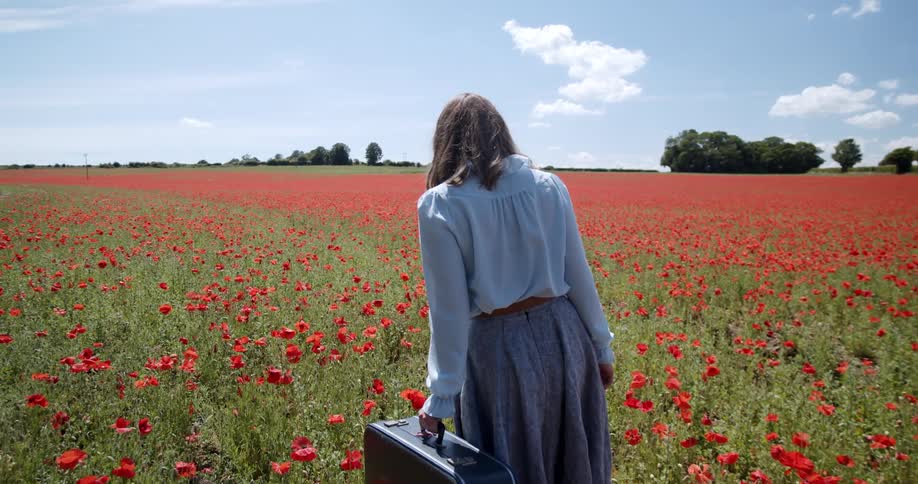 A woman in old fashioned clothing carries luggage through a vast field of red flowers