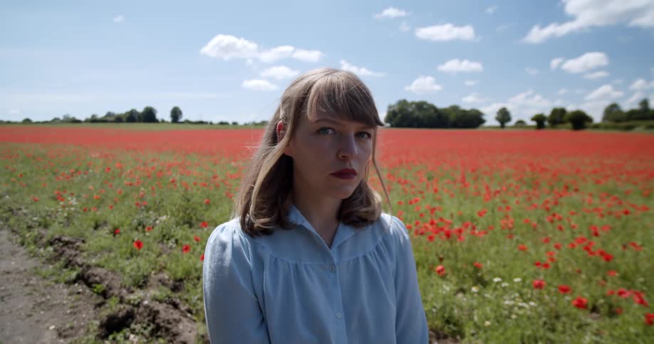 Blonde woman standing in front of a prairie filled with red wild flowers looking at the camera