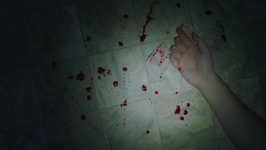 A hand lays in the ground surrounded by drops of fresh red blood in a dark room