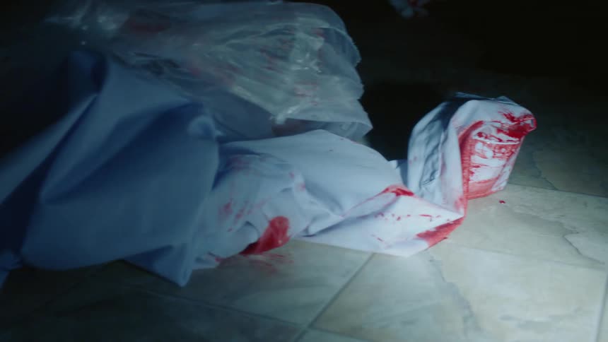Clothes drenched in fresh red blood laying in the ground in a dimly lit room