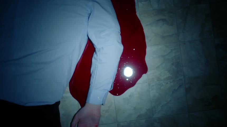 Close up of a mans body laying in the ground over a pool of fresh red blood in a dimly lit room