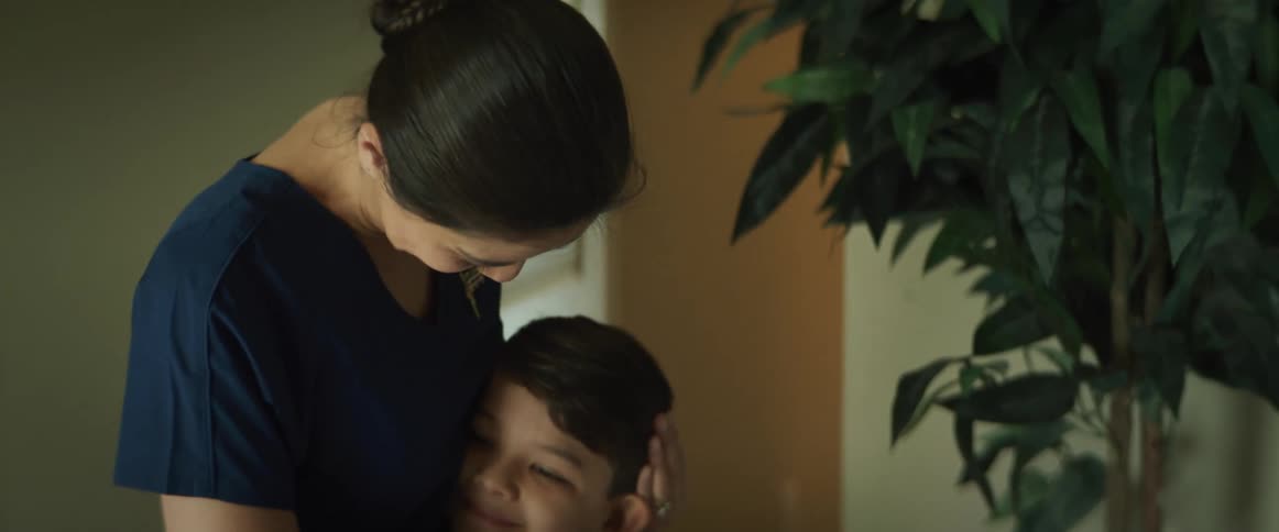 A nurse in scrubs hugs her son before going to work. They walk out of her room together.
