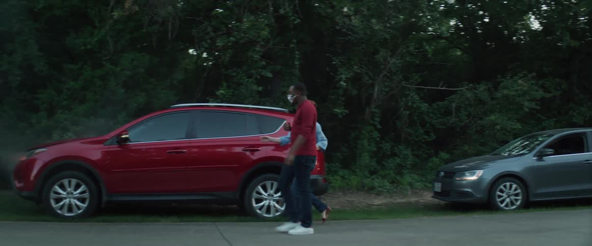 A woman stopped at the side of the road with smoke coming out of her car talks on the phone as a man stops and gets out and goes and open the hood to look