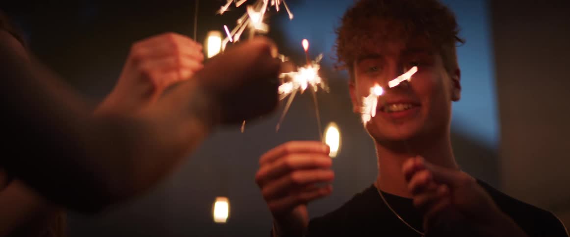 A group of laughing young men and women hold sparklers together while standing in a circle outdoors at night