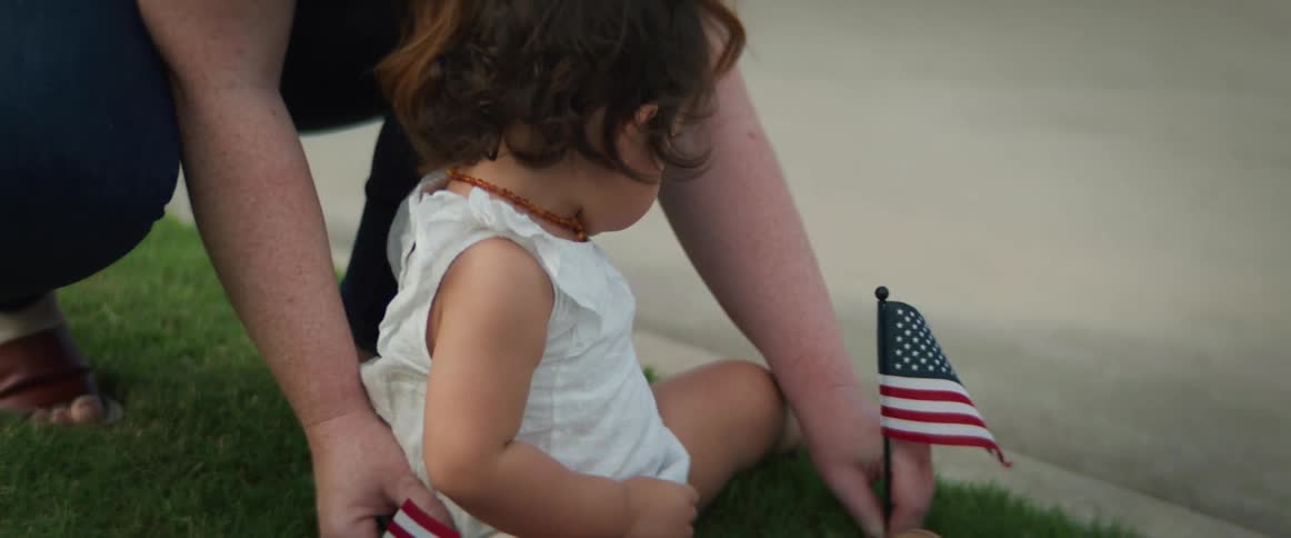 A woman puts a little flag in the grass and her baby pulls it out