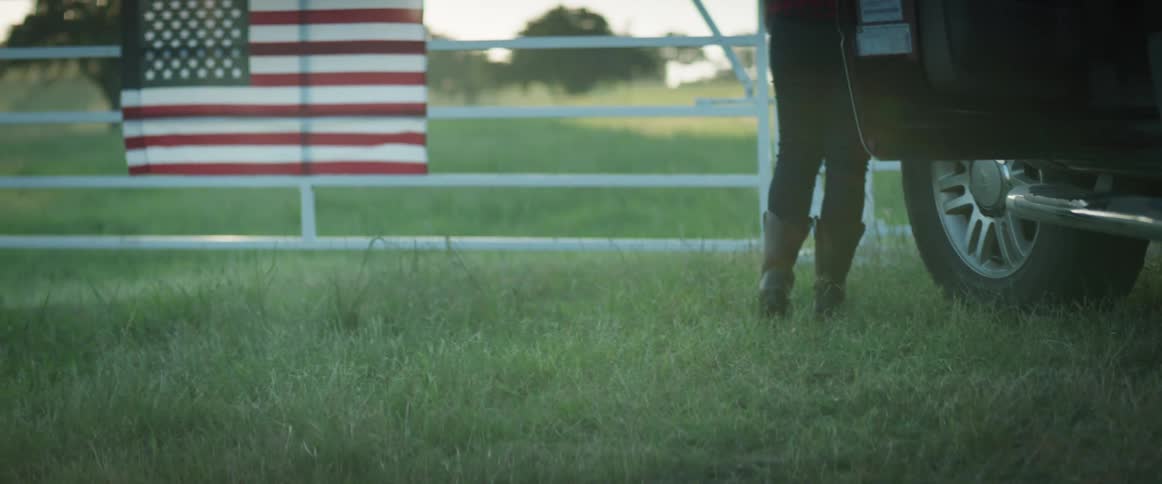 A woman exits a vehicle and goes to rest against a gate next to an american flag