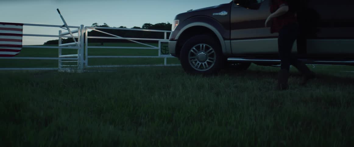 A woman drives up to a fence on a farm and gets out of her vehicle and walks over to the fence and looks across the land