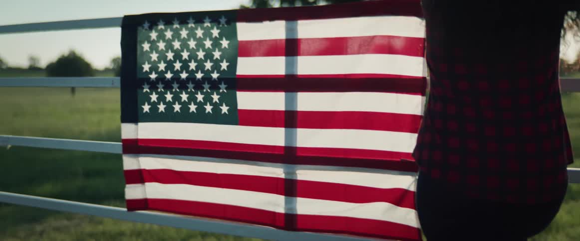 A woman leans against a fence with an american flag on it and looks at the land in front of her