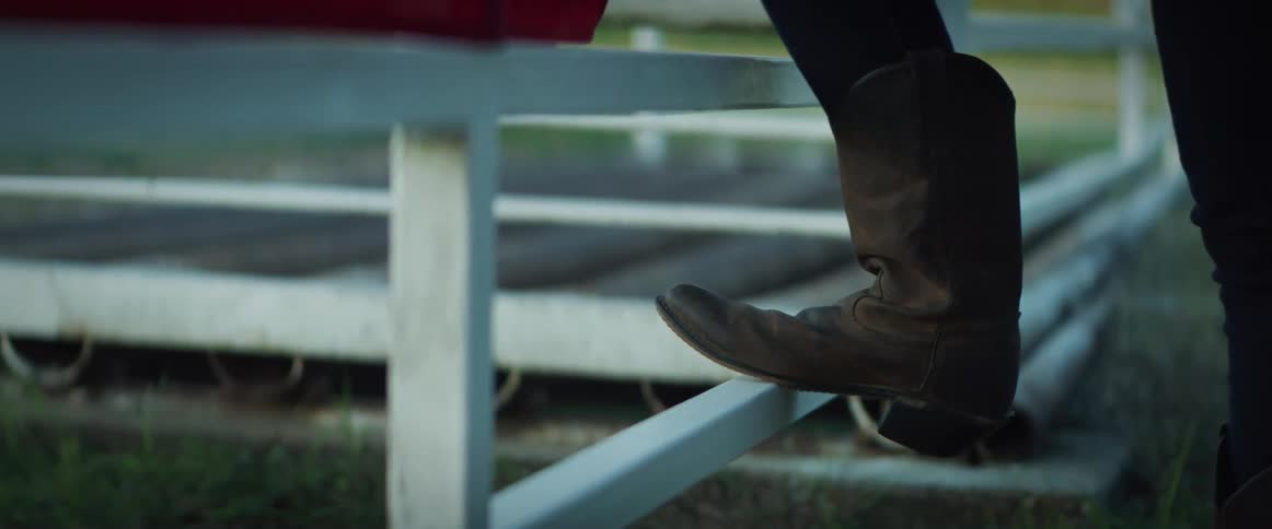 A cowboy walks up to a metal fence and rests their foot on it