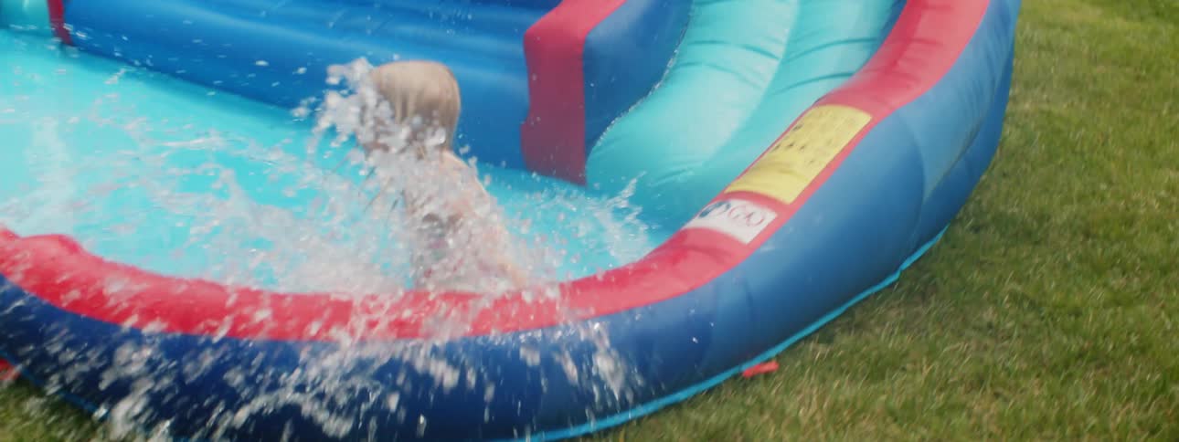 A little girl in swimwear slides down an inflatable water slide in a yard on a sunny day