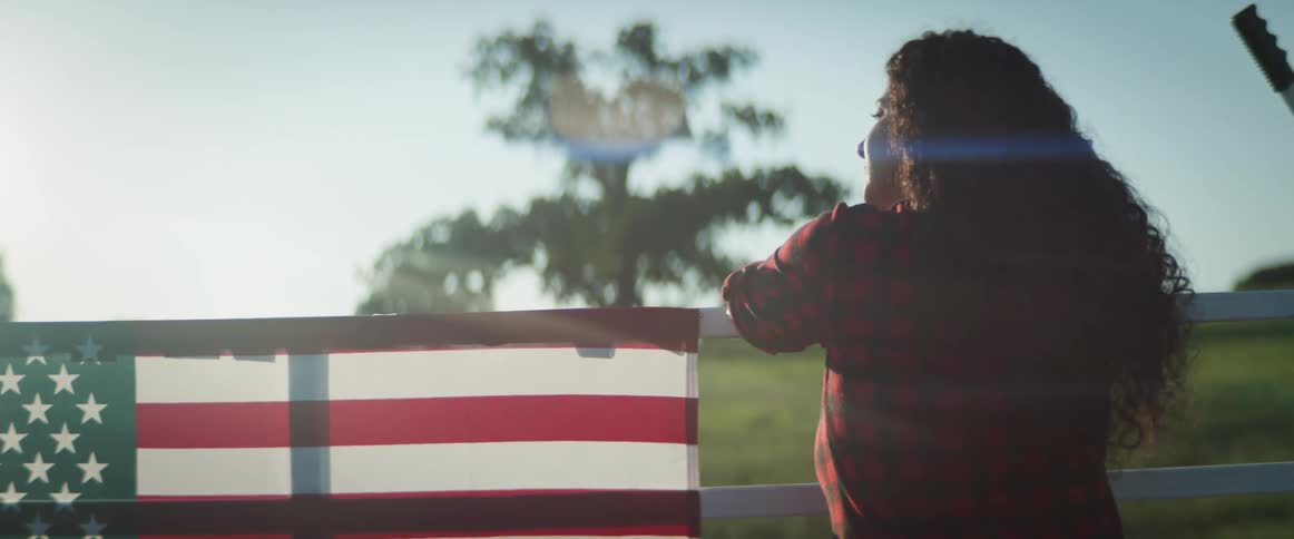 Young woman reclines on a fence with an american flag in a field at sunset