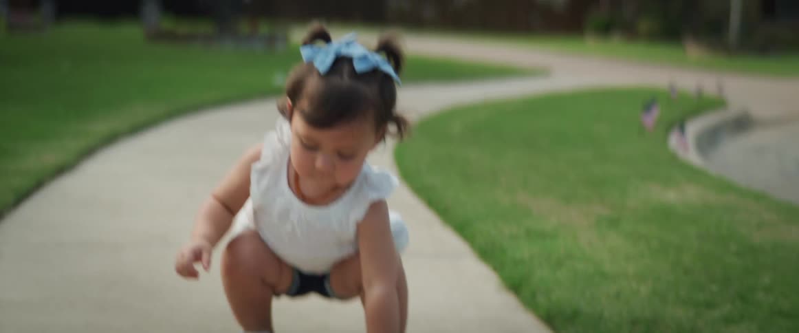 A young toddler girl holds a small flag drops it and bends down to the sidewalk to pick it up