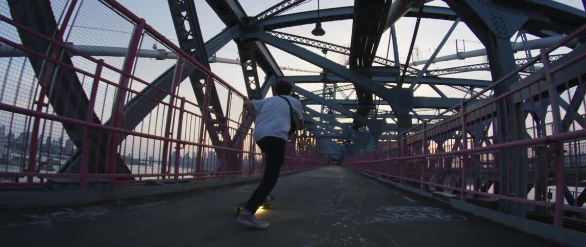 A person rides a lighted skateboard on a large pedestrian bridge over a river