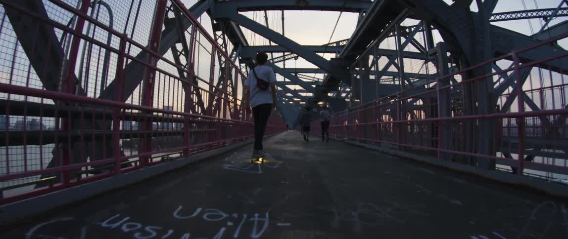 A teenager rides on a skateboard with under lights near people walking across a bridge