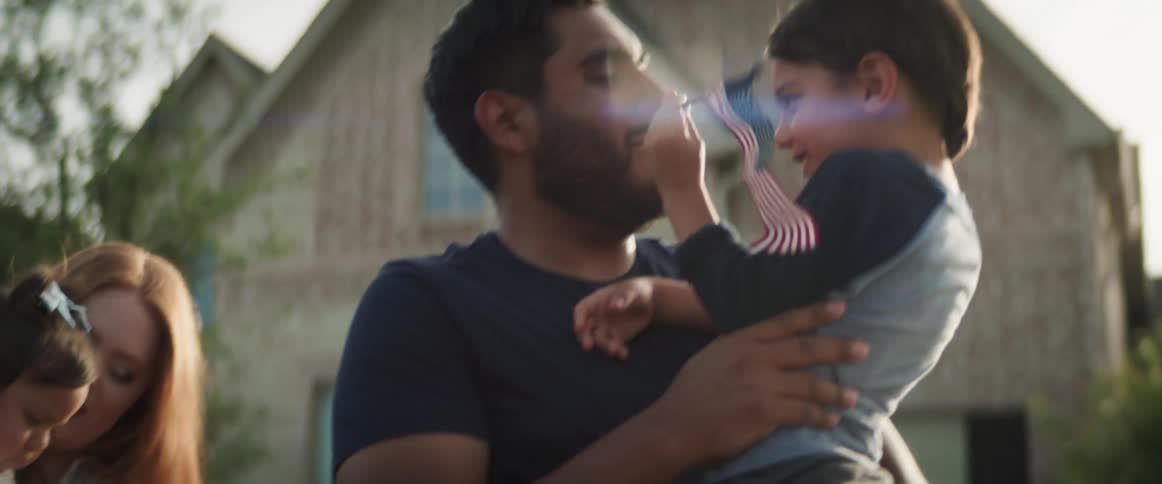 A family with two small children stands outside waving small american flags together