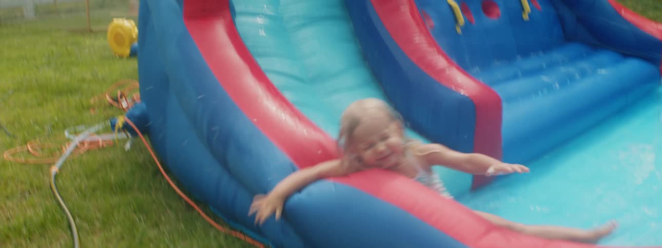Little girl in a swimsuit slides down an inflatable pool playground water slide on the backyard