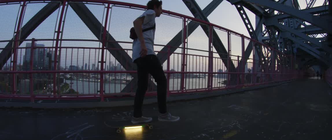 A teenage boys skateboards across a bridge with the water and city skyline in the distance