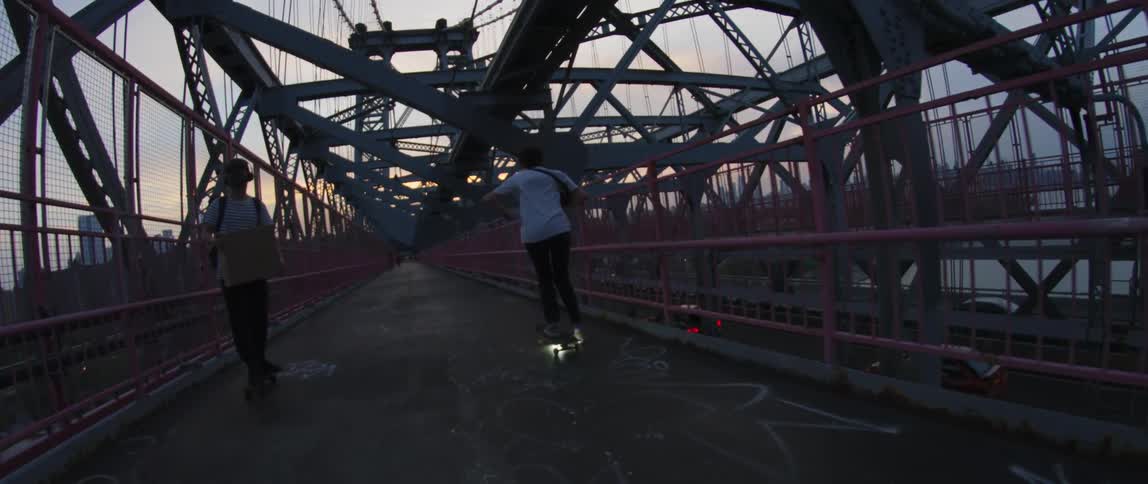 Two skateboarders pass each other on bridge crossing river overlooking highway traffic