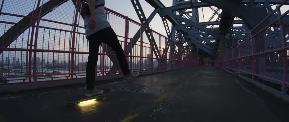 A man rides a skateboard with a glowing flourescent light mounted on the bottom across a bridge over a city bay