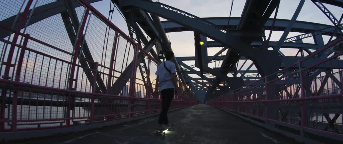 Person skateboards over bridge crossing river toward city on the horizon