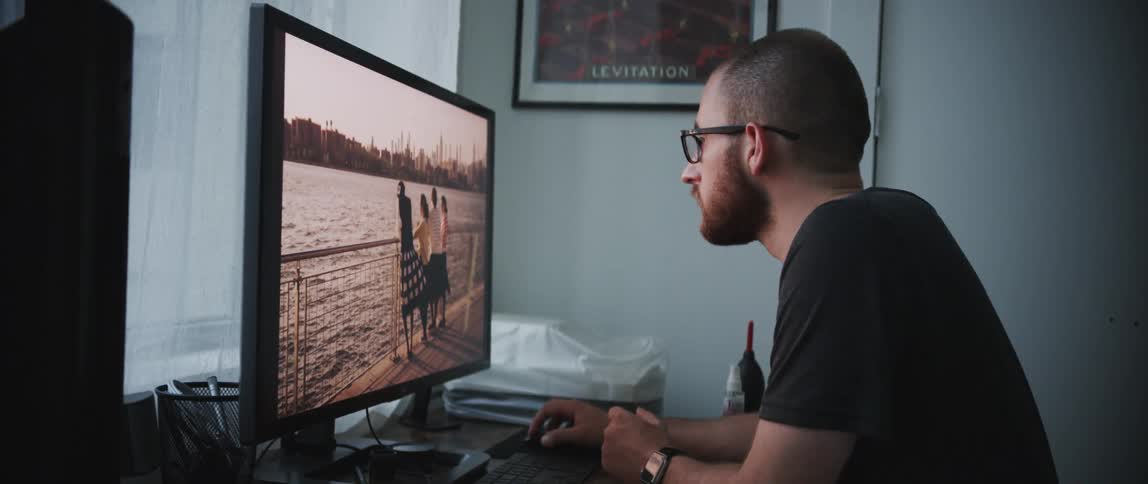 A man with glasses and a beard focuses on a computer screen as he edits a photo of some people