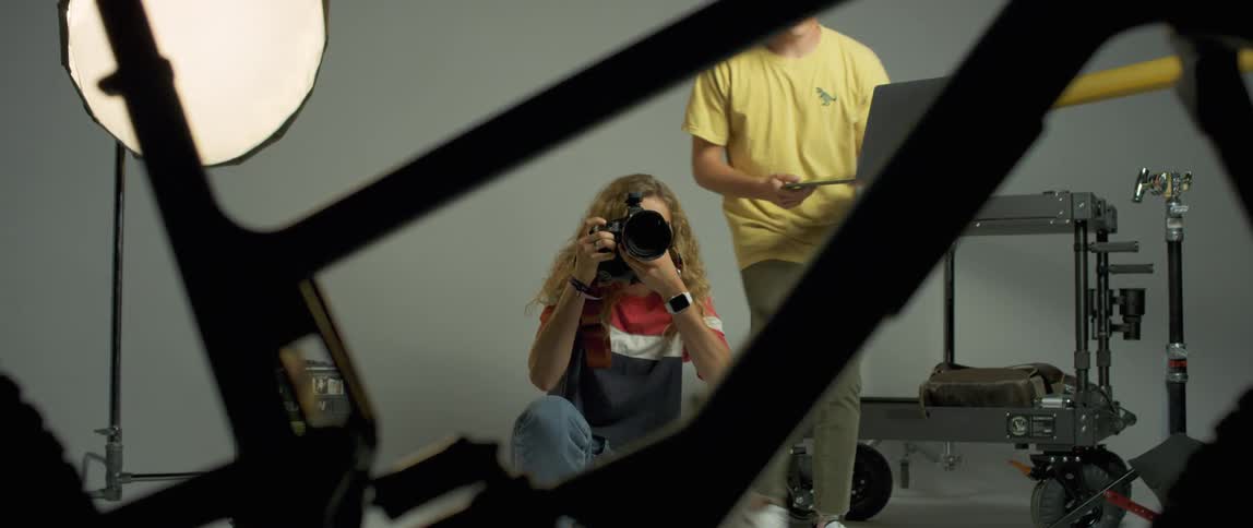 A man carries a laptop to show it to a female photographer taking photographs of a bicycle in a studio