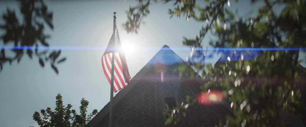 The american flag attached to a pole by a house flutters in the wind under the sun