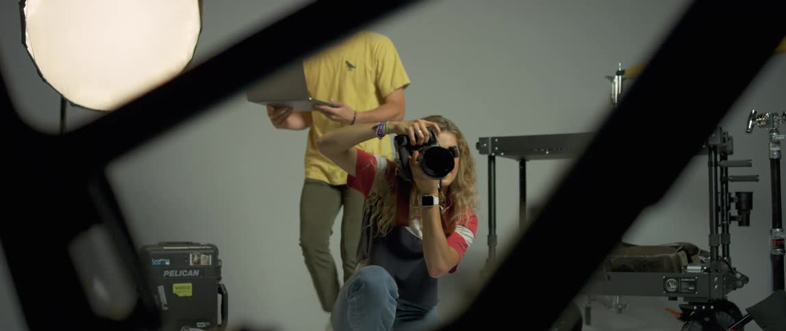 A female photographer kneels to photograph a bicycle as a man retrieves a laptop from a cart and approaches her from behind
