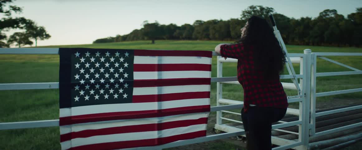 American flag hangs on a fence while a woman walks up to the fence and leans on it while looking out at a field