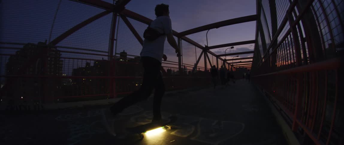 Man rides skateboard along bridge passing pedestrians walking the other way with city buildings showing on both sides of bridge