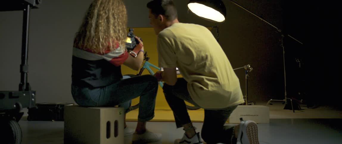 A photographer takes photos of a bike and then a young man shows her pictures on a laptop and looks at her camera