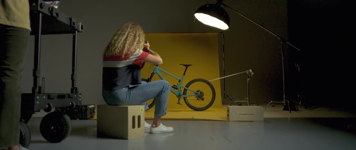 Female photographer sitting on a box takes pictures of a turquoise bicycle in a studio