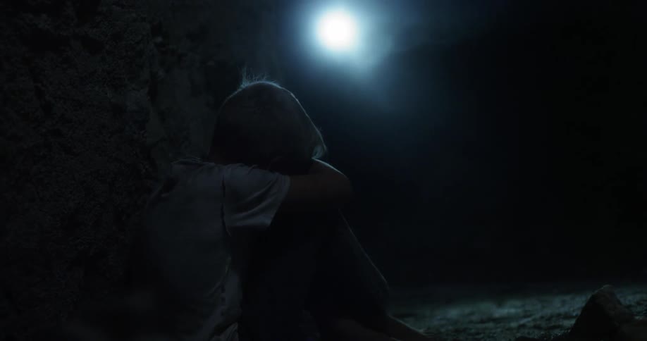 A young boy sitting with head down in a dark place as a flashlight approaches